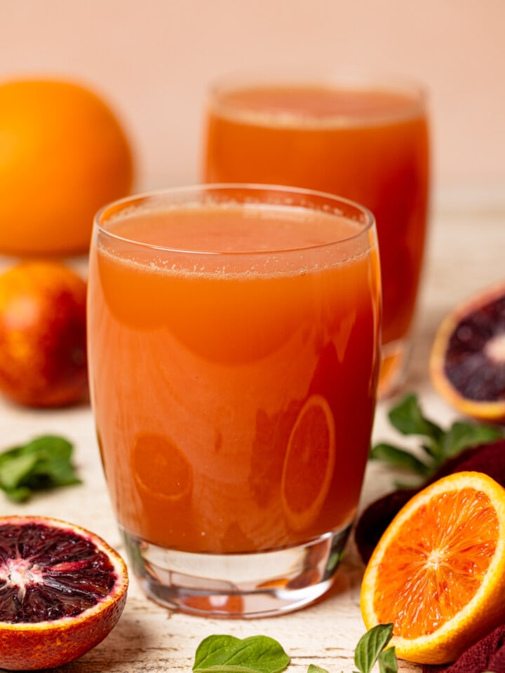 Two glasses of juices on a white table with blood oranges and herbs.