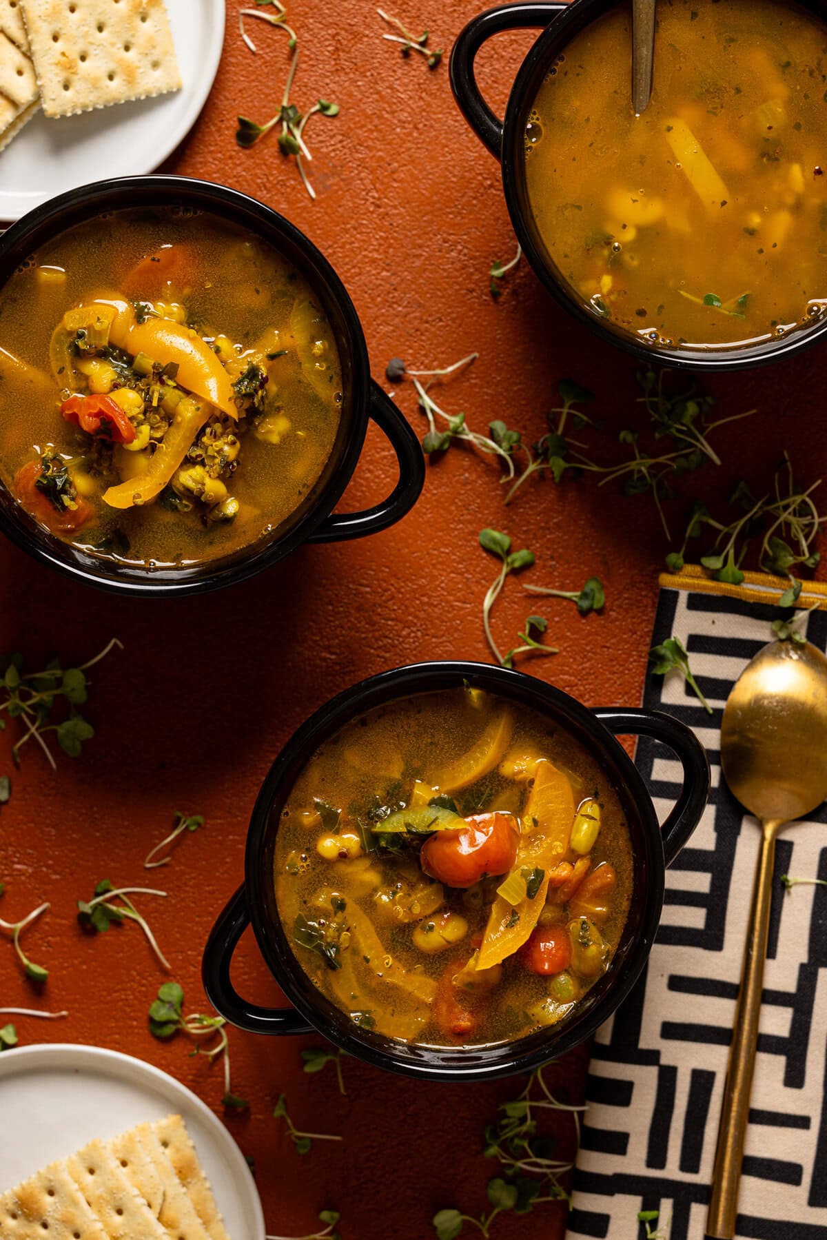 Three bowls of soup on a burnt orange table with a spoon and crackers.