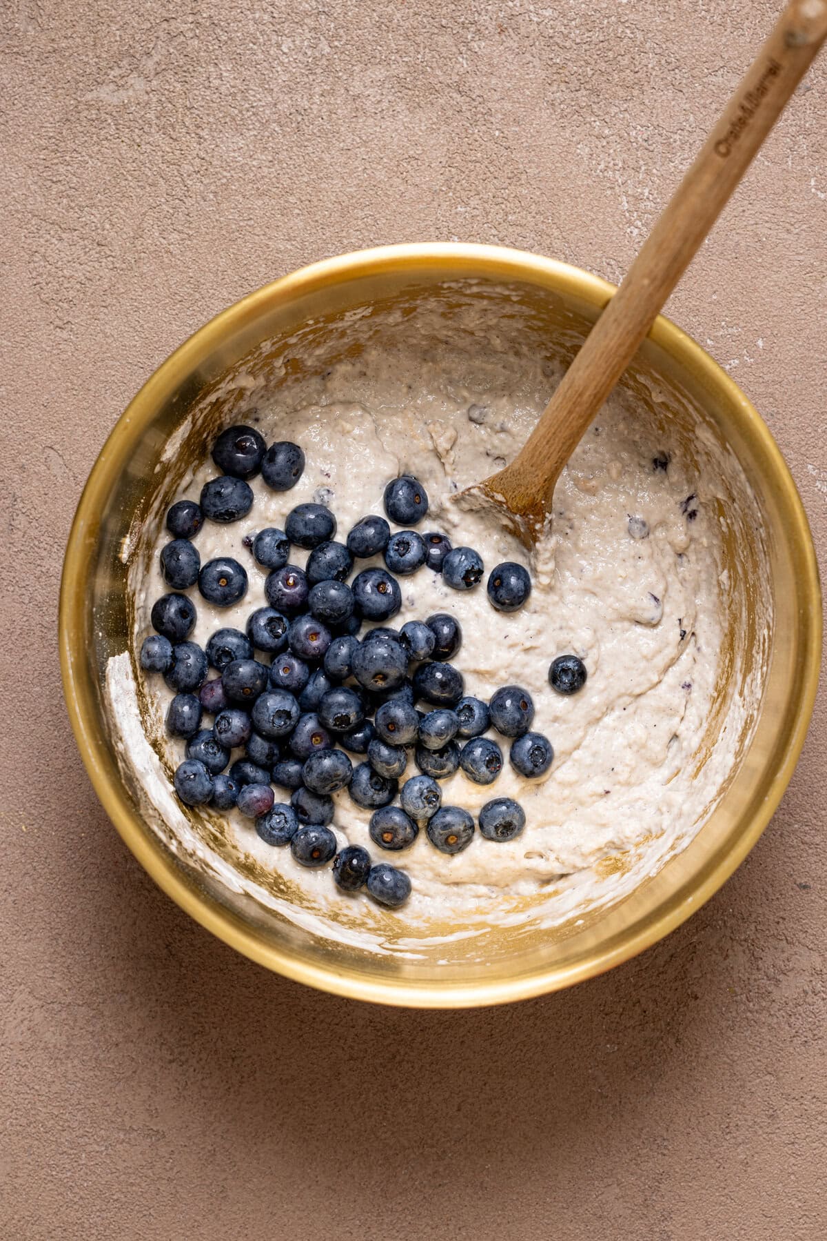 Blueberries atop batter in a bowl with a wooden spoon.