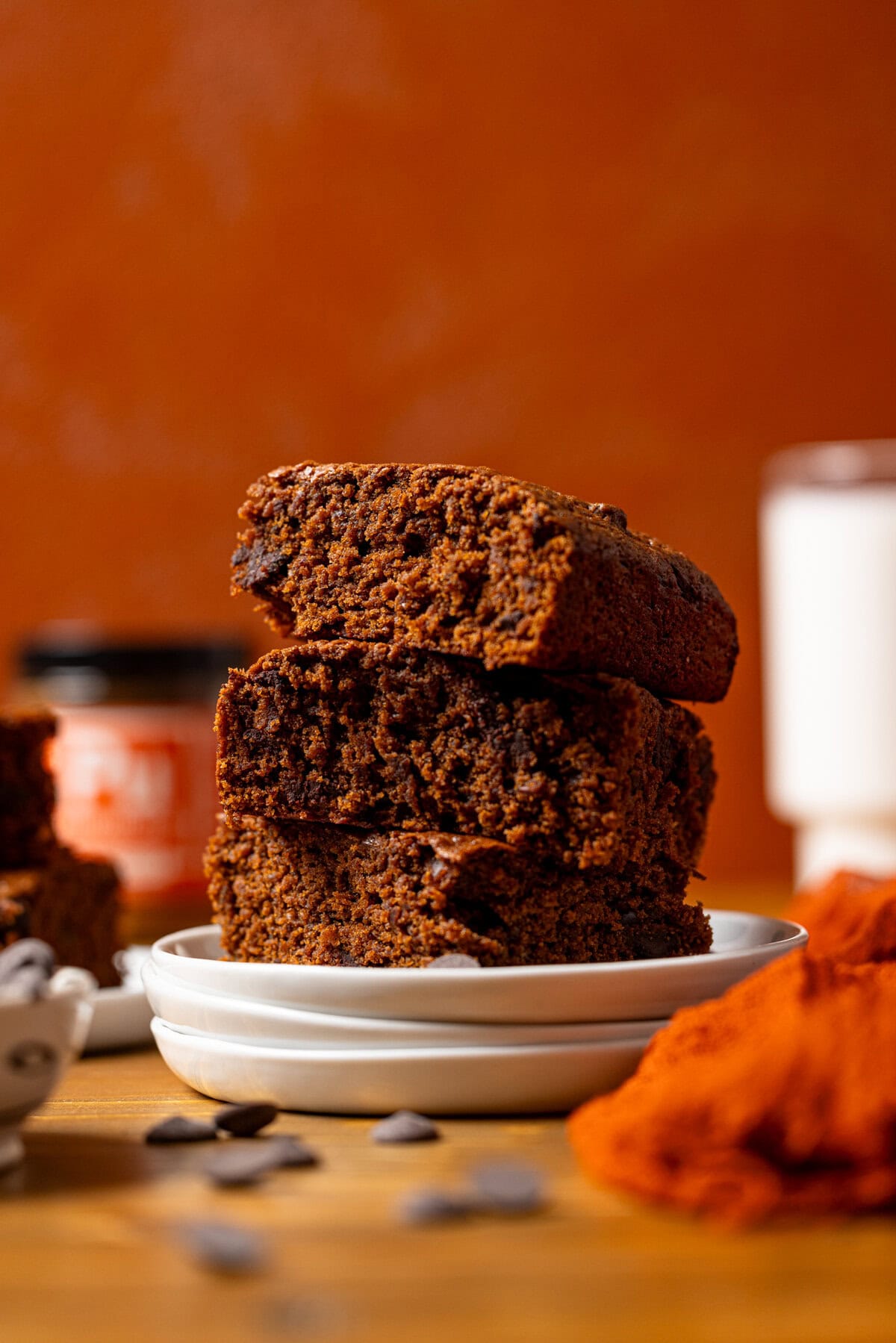 Stack of sweet potato brownies on white plates with a glass of milk, red napkin, and chocolate chips.