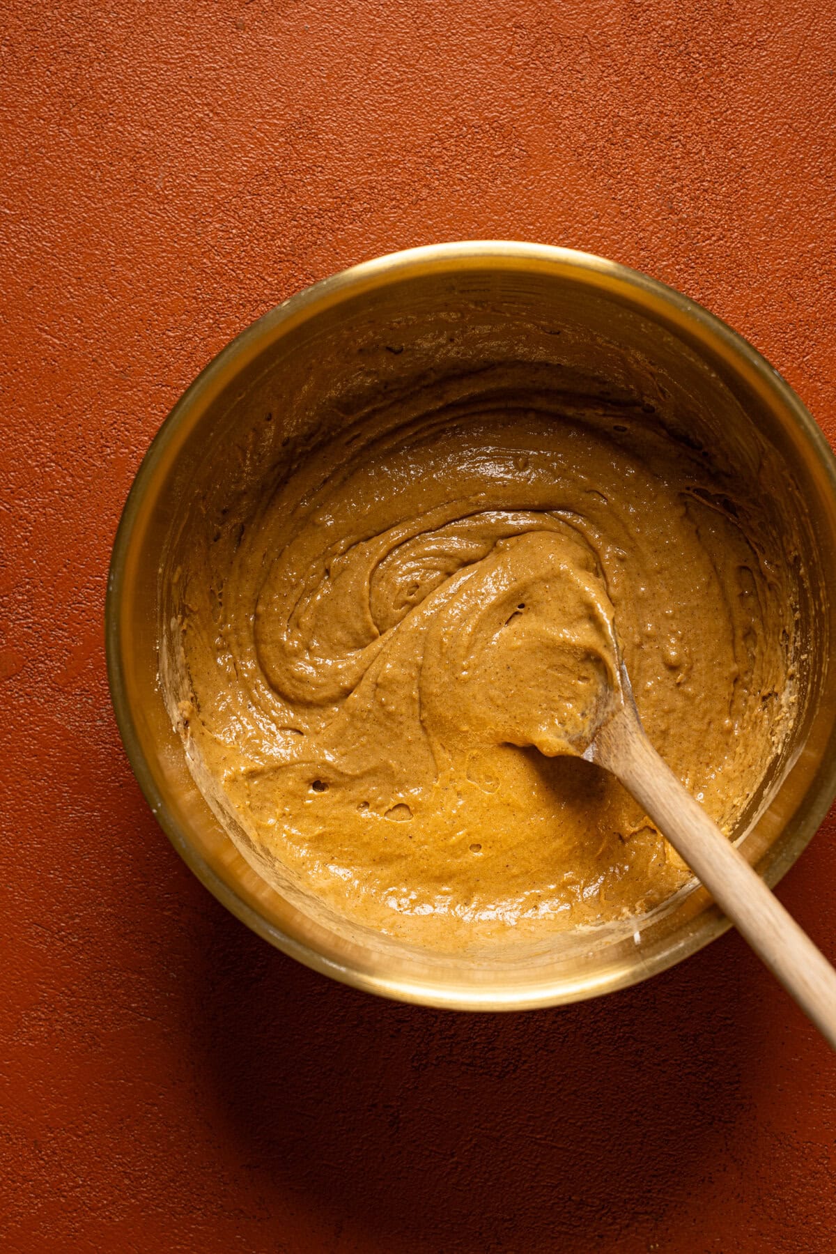 Gingerbread muffin batter in a gold bowl with a wooden spoon.