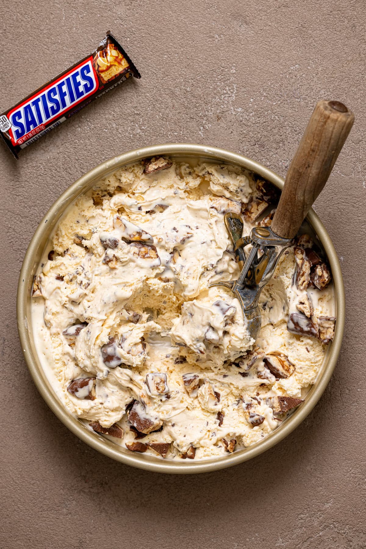 Snickers ice cream in a round metal pan on a greyish brown table with a scoop and candy bar.