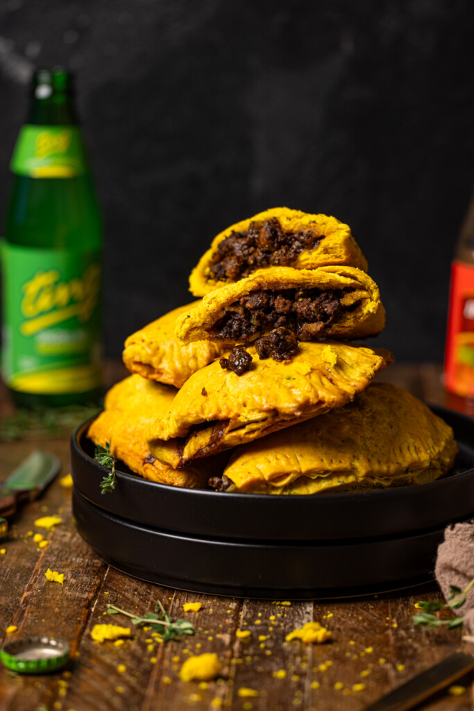 Up close shot of Jamaican beef patties stacked on black plates with a drink, hot pepper sauce, and knife.