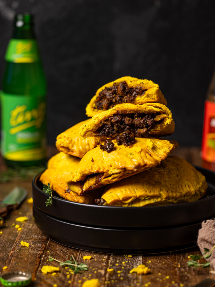 Up close shot of Jamaican beef patties stacked on black plates with a drink, hot pepper sauce, and knife.