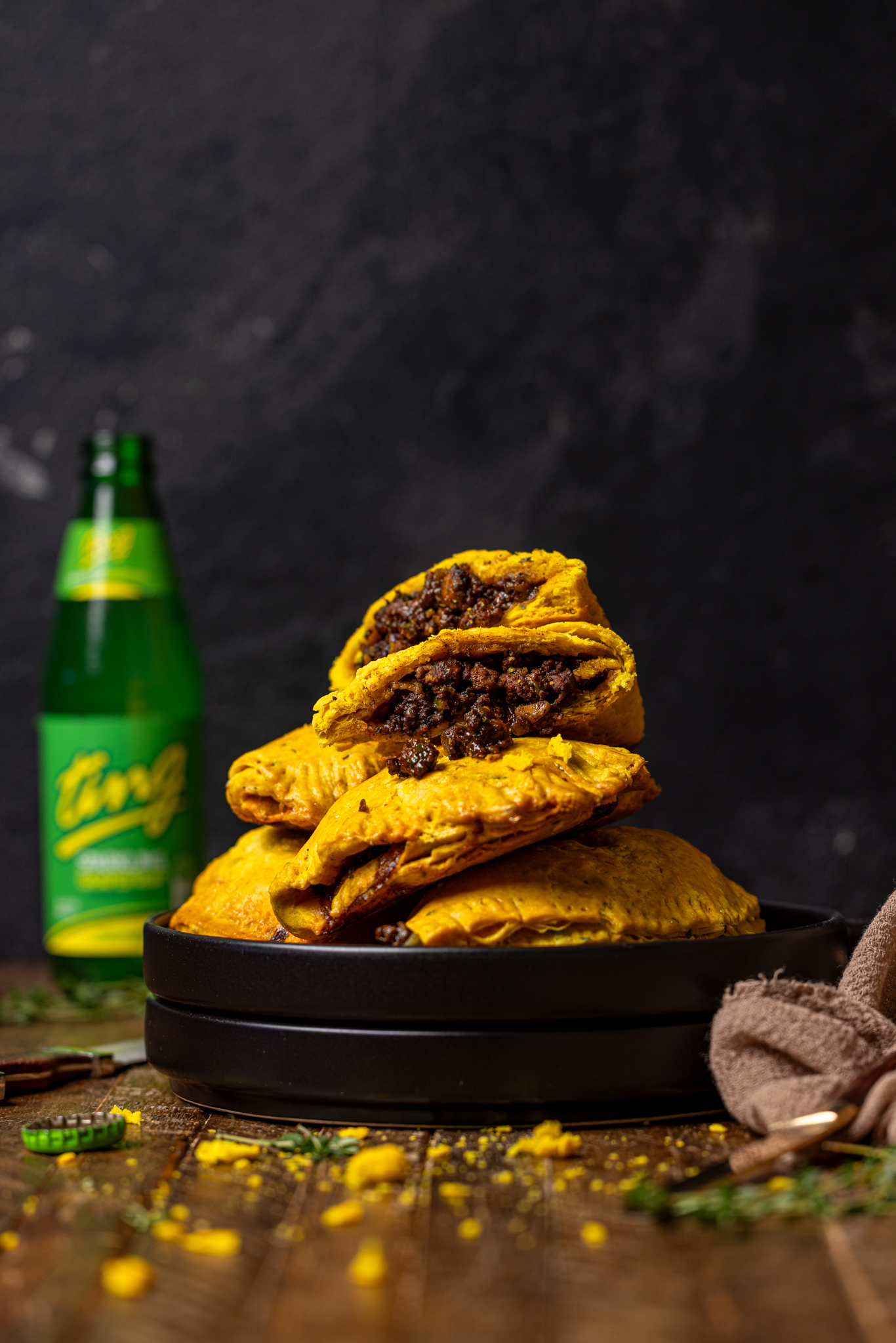 Image of Jamaican beef patties stacked on black plates with a bottle of ting.