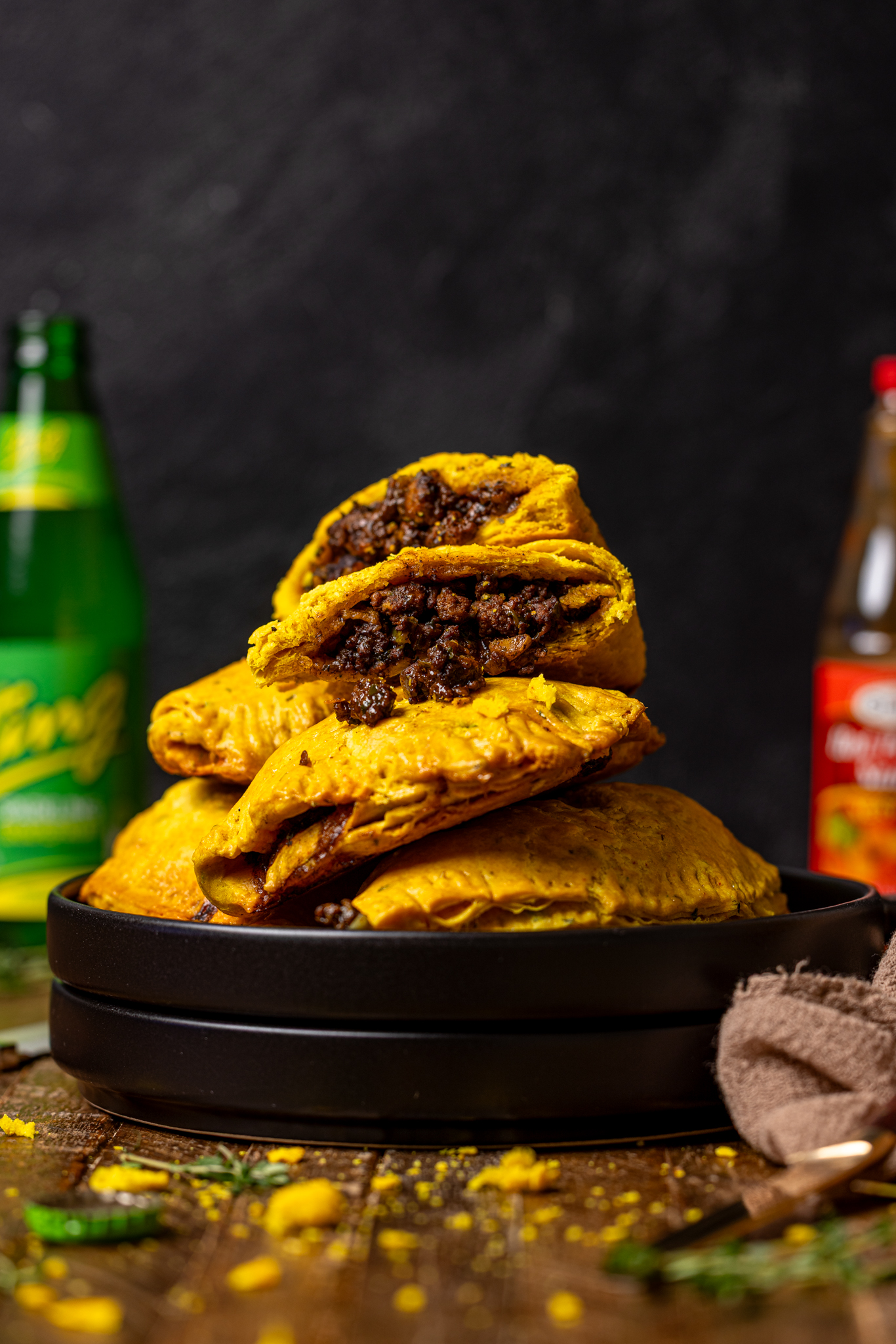 Image of stacked Jamaican beef patties with a bottle of ting and hot pepper sauce.