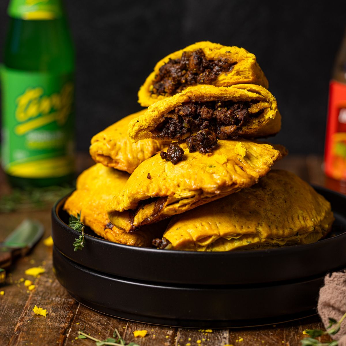 Up close shot of Jamaican beef patties stacked on black plates with a drink, hot pepper sauce, and knife.