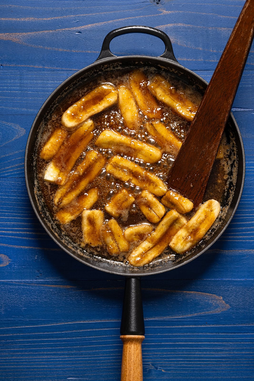 Caramelized bananas in a black skillet with a wooden spoon.