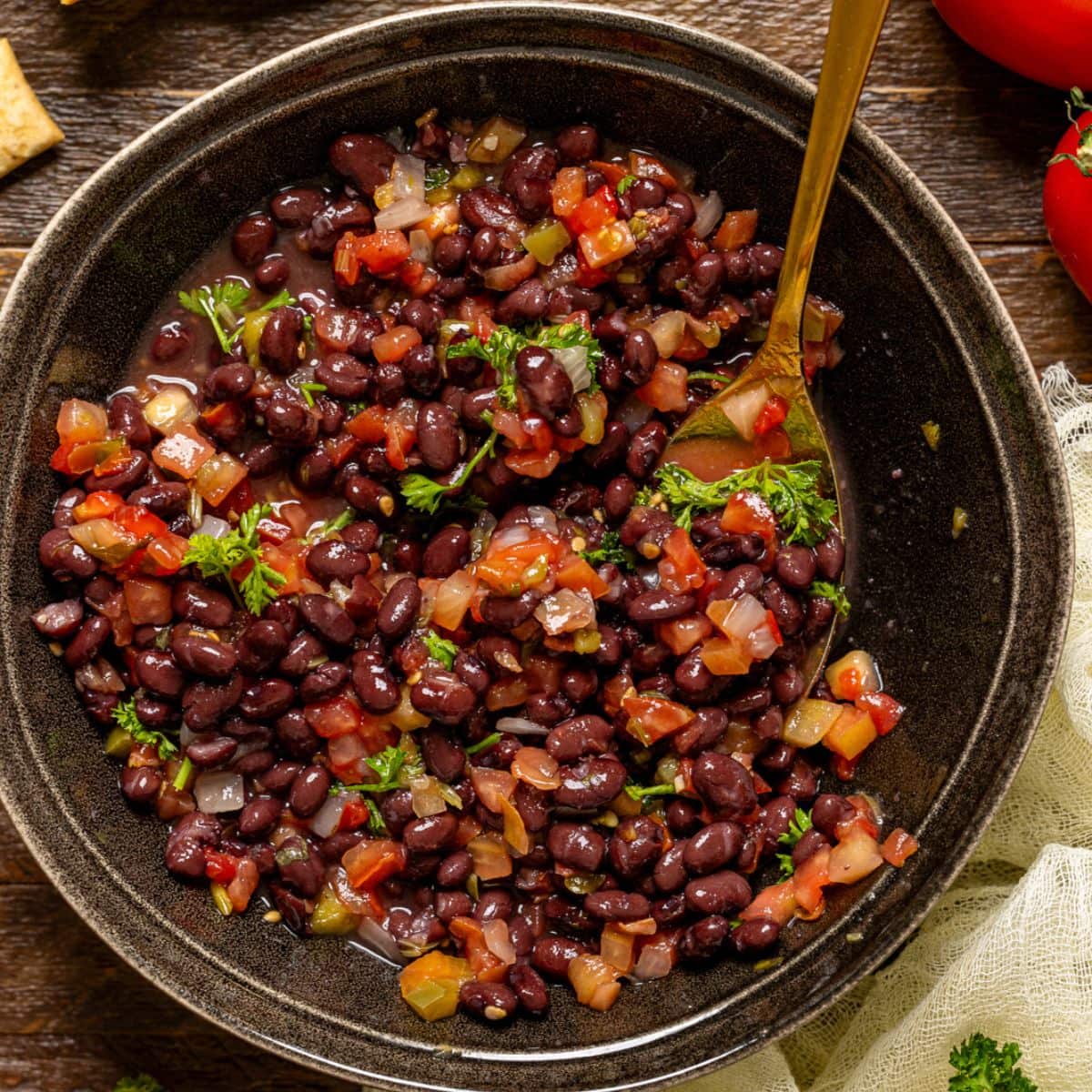 Black bean salad in a bowl with a gold spoon, crackers, and tomatoes.