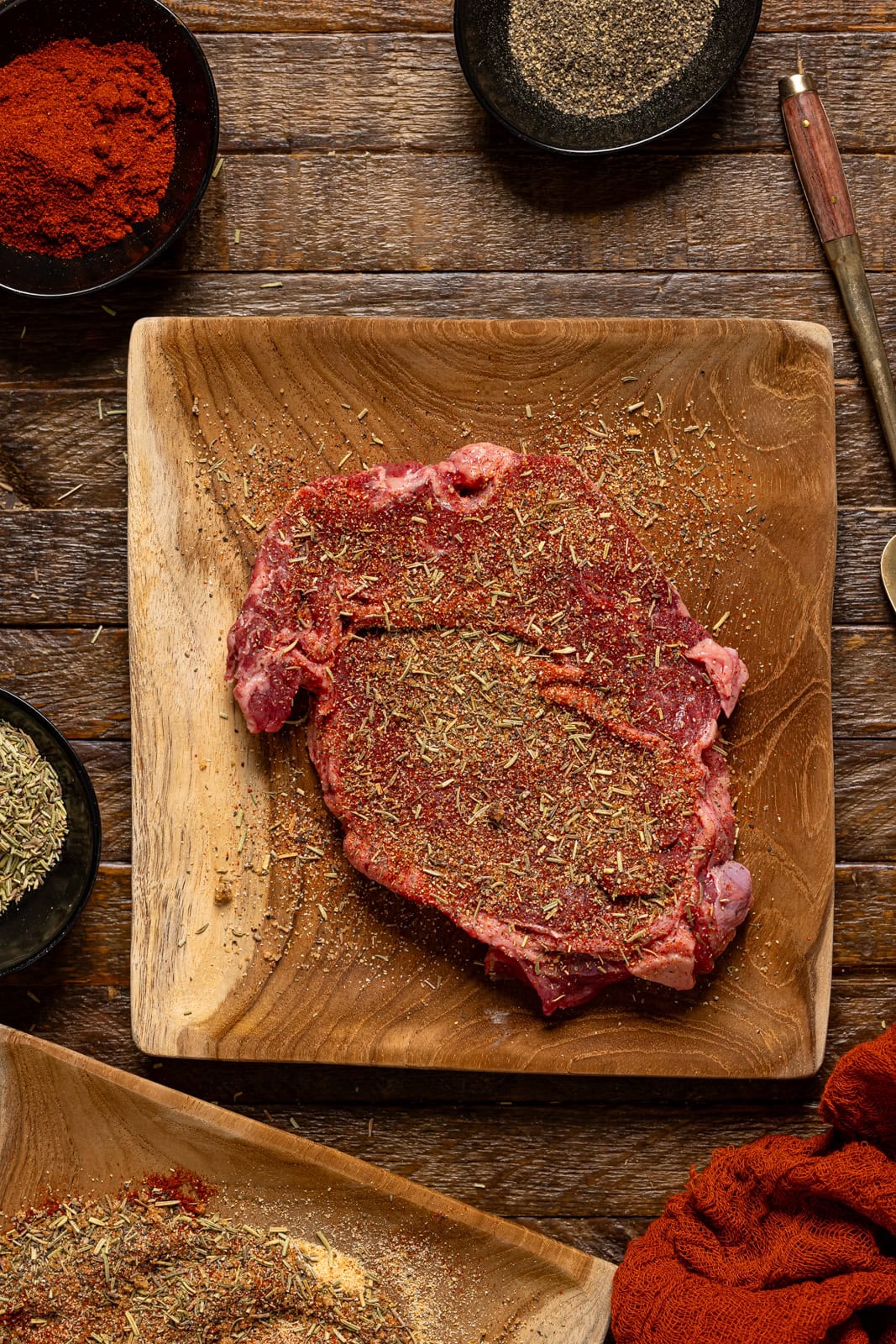 Seasoned steak on a brown wood plate with seasonings on the side.