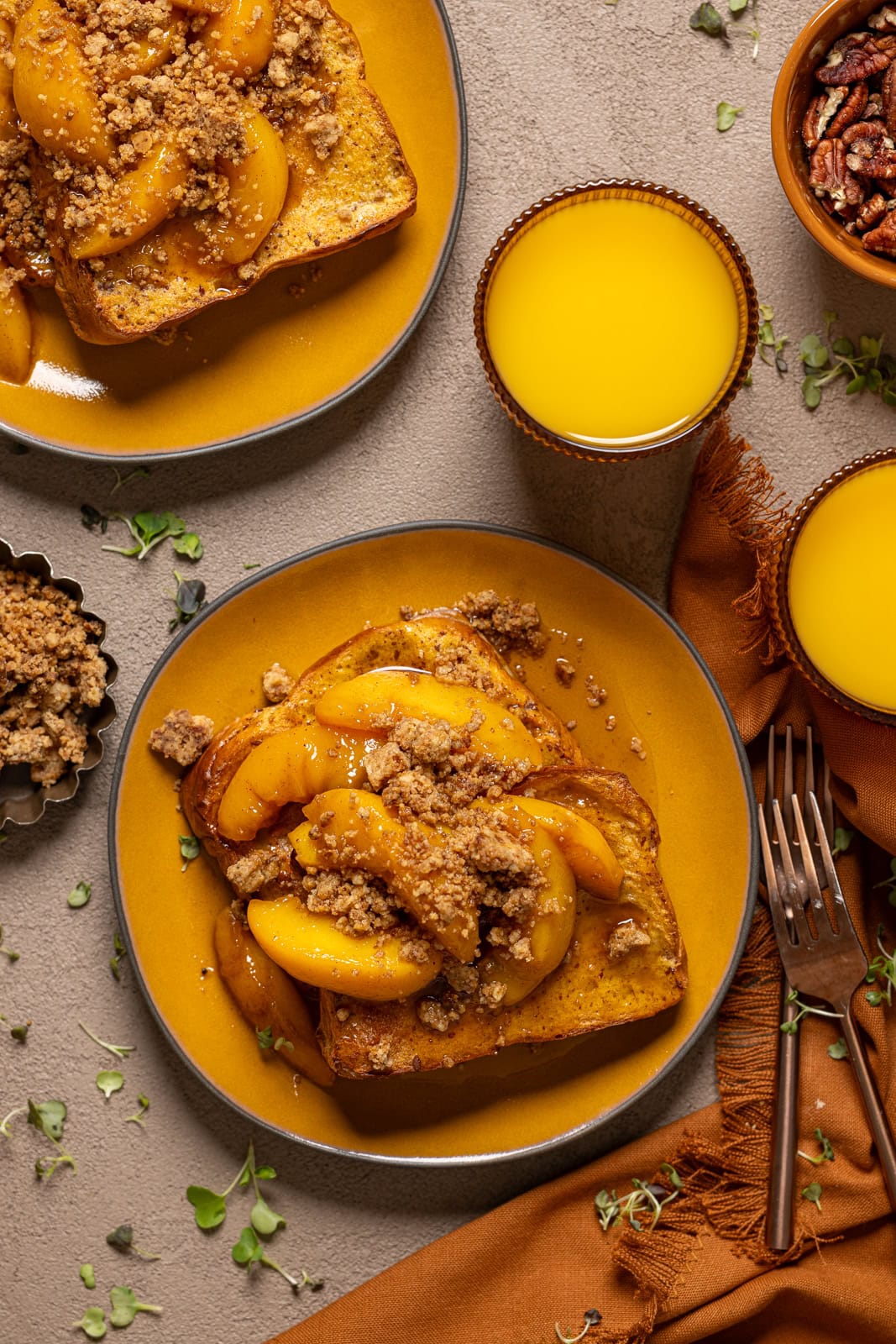 Two plates of french toast with glasses of orange juice with pecans and forks.