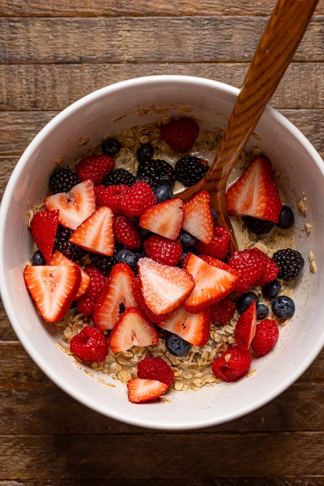 Fresh berries and ingredients in a white bowl with a wooden spoon.