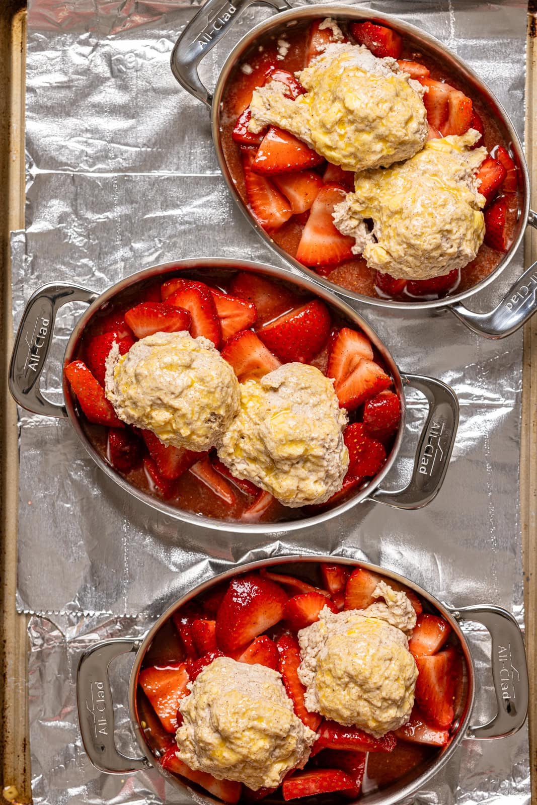 Strawberry cobblers assembled in three metal bakeware dishes on a baking sheet with foil.