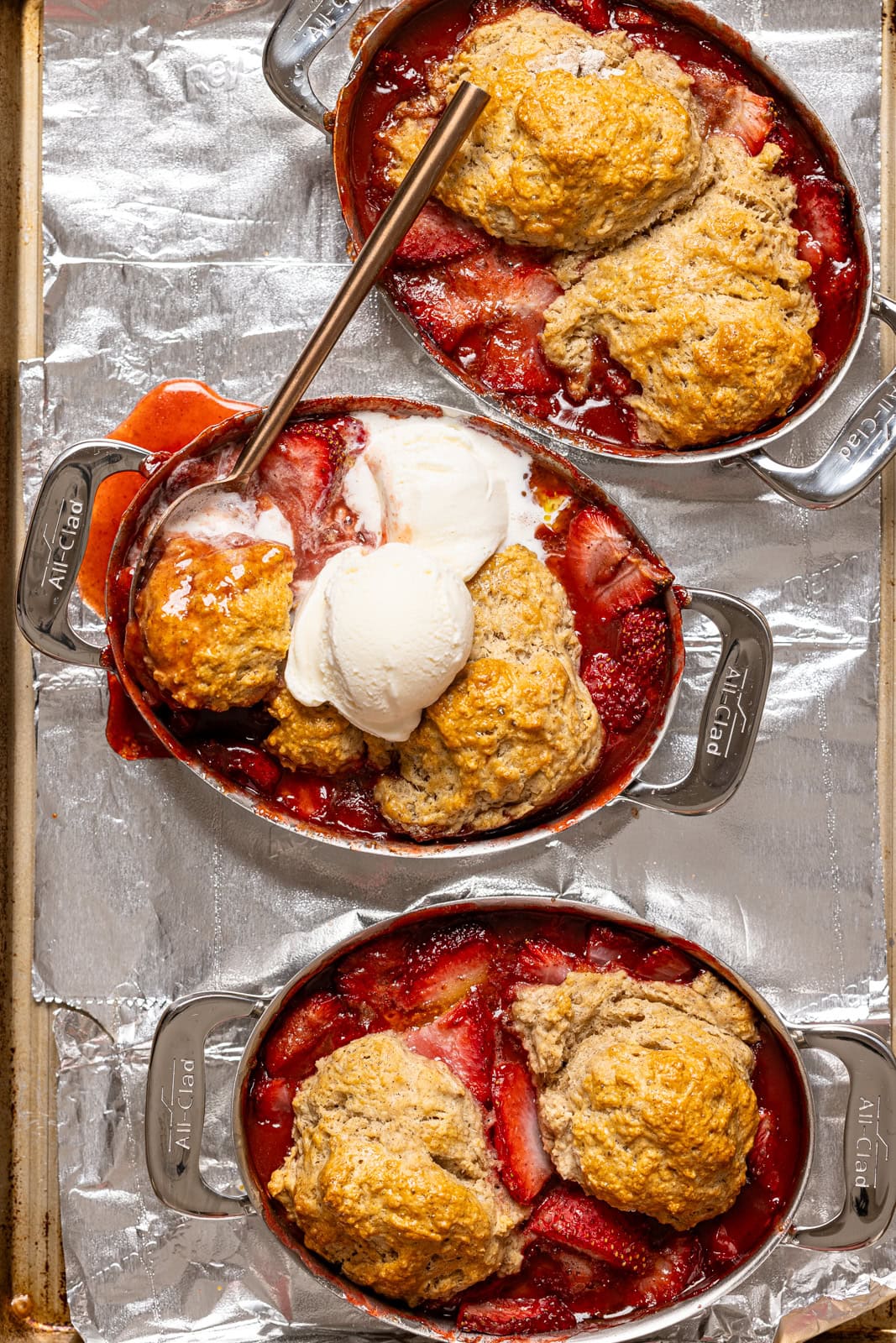Baked cobblers on a baking sheet with foil and ice cream and a spoon.
