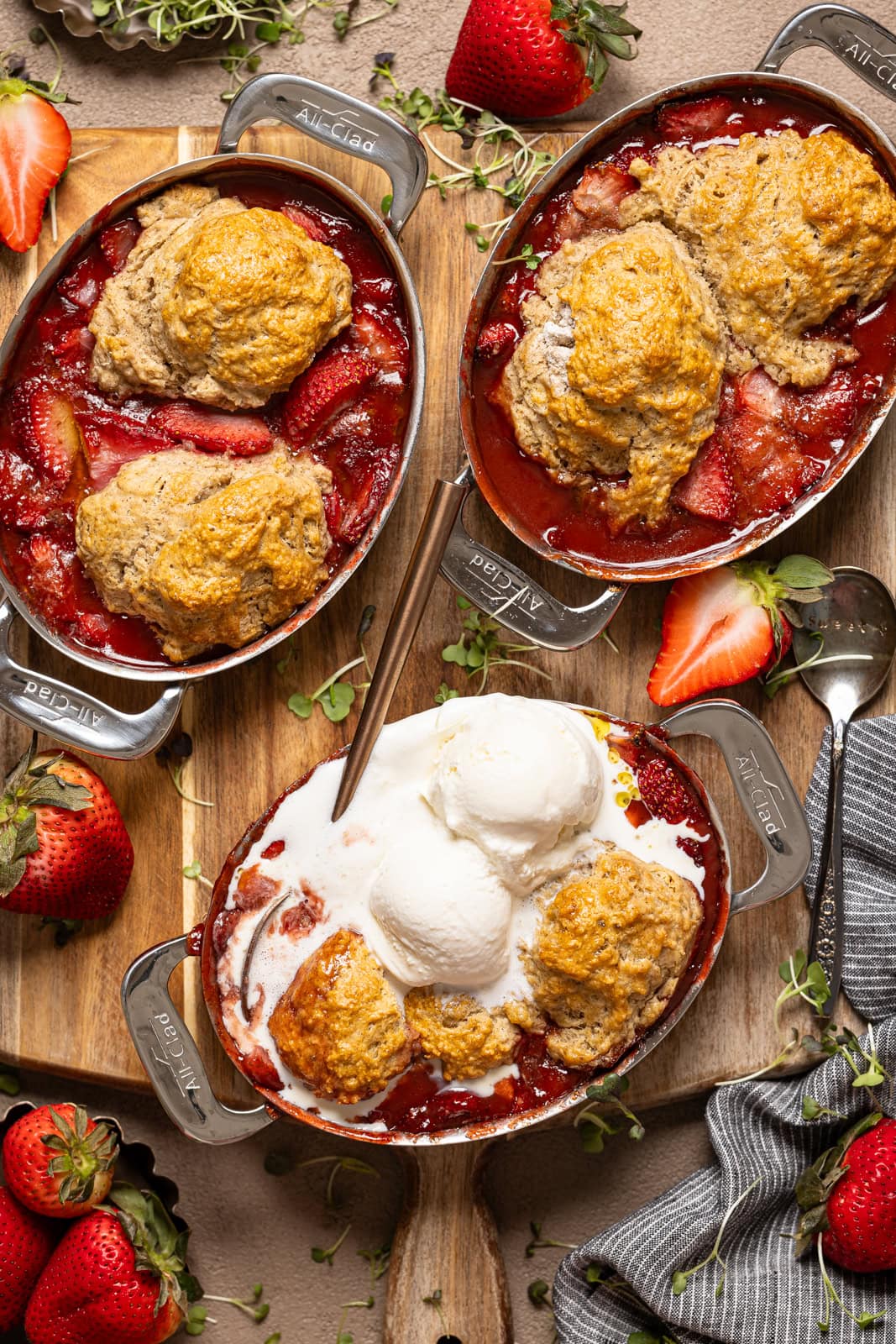 Strawberry cobbler in three metal bakeware with spoons and strawberries. 