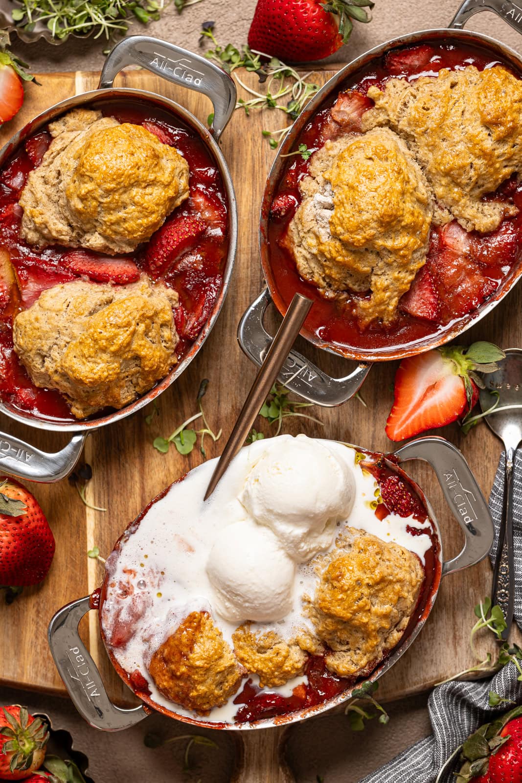 Three metal bakeware dishes with strawberry cobbler and a scoop of ice cream and spoons.