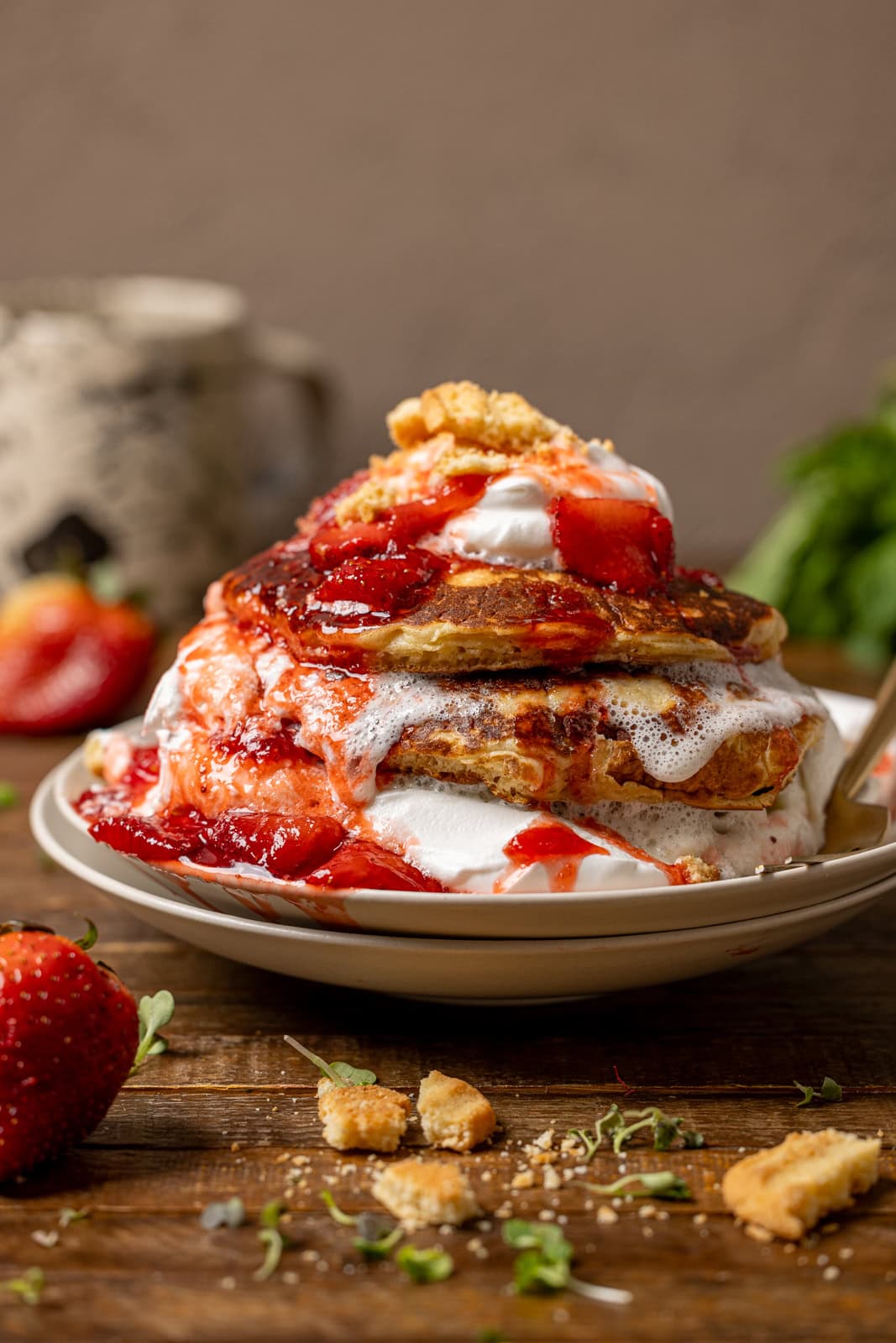 Pancakes on a stack of plates with whipped ream, strawberries, crushed cookies on a brown wood table.