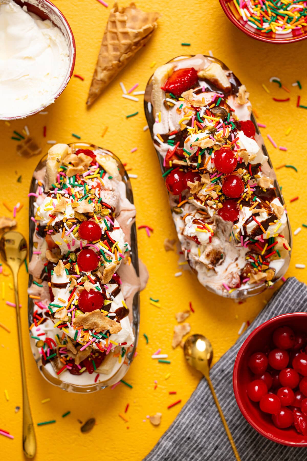 Overhead view of banana split on a yellow table with cherries, spoons, and cones.
