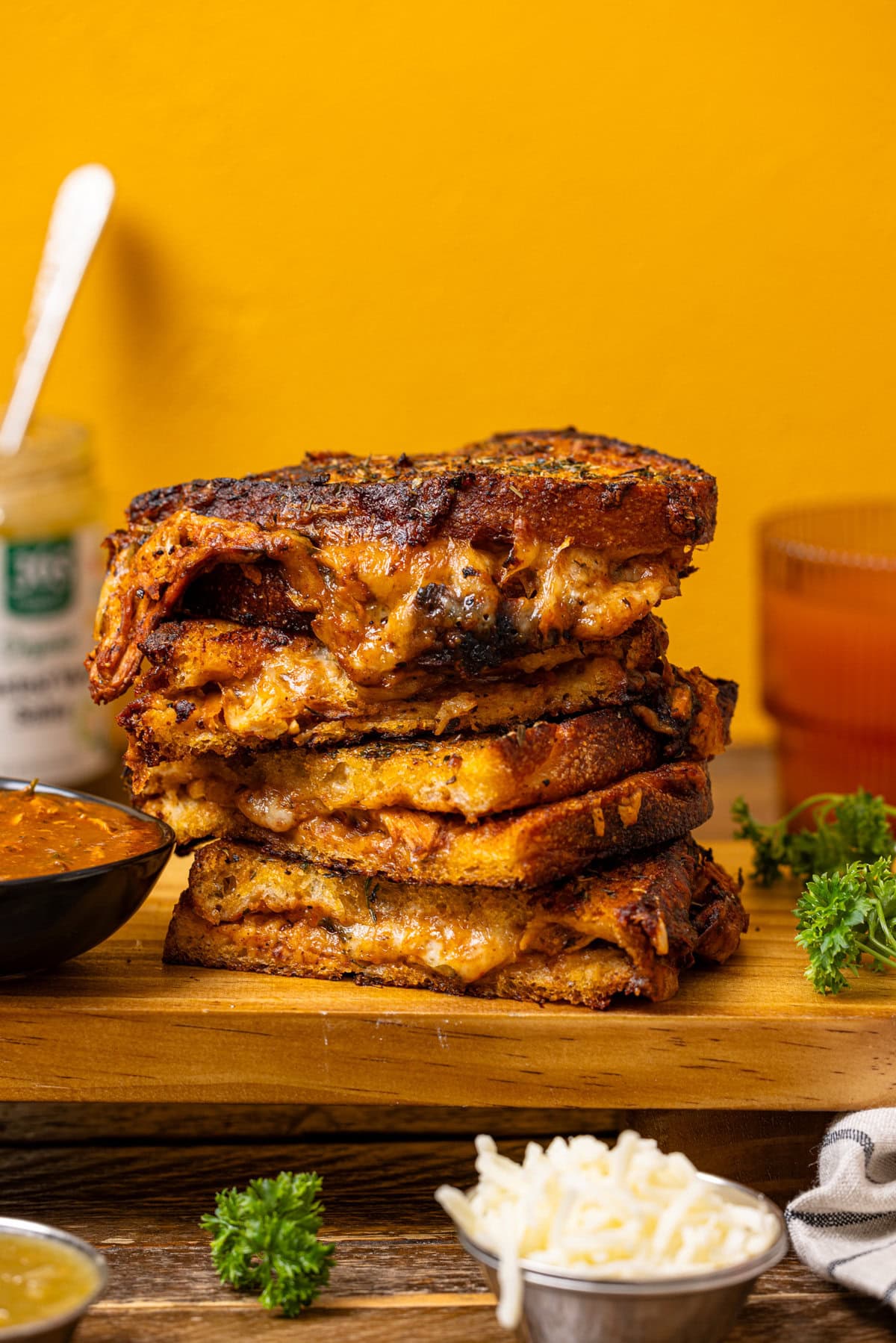 Up close shot of stacked grilled cheese on a wooden tray with cheese, a drink, and sauce. 