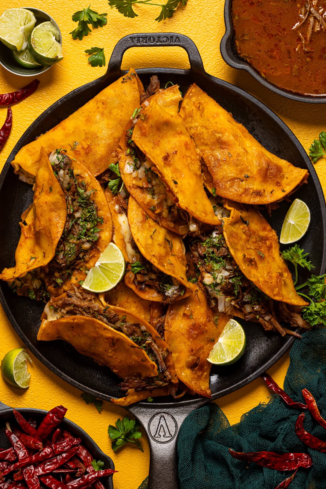 Birria tacos on a black skillet platter with lime, sauce, and peppers on a yellow table.