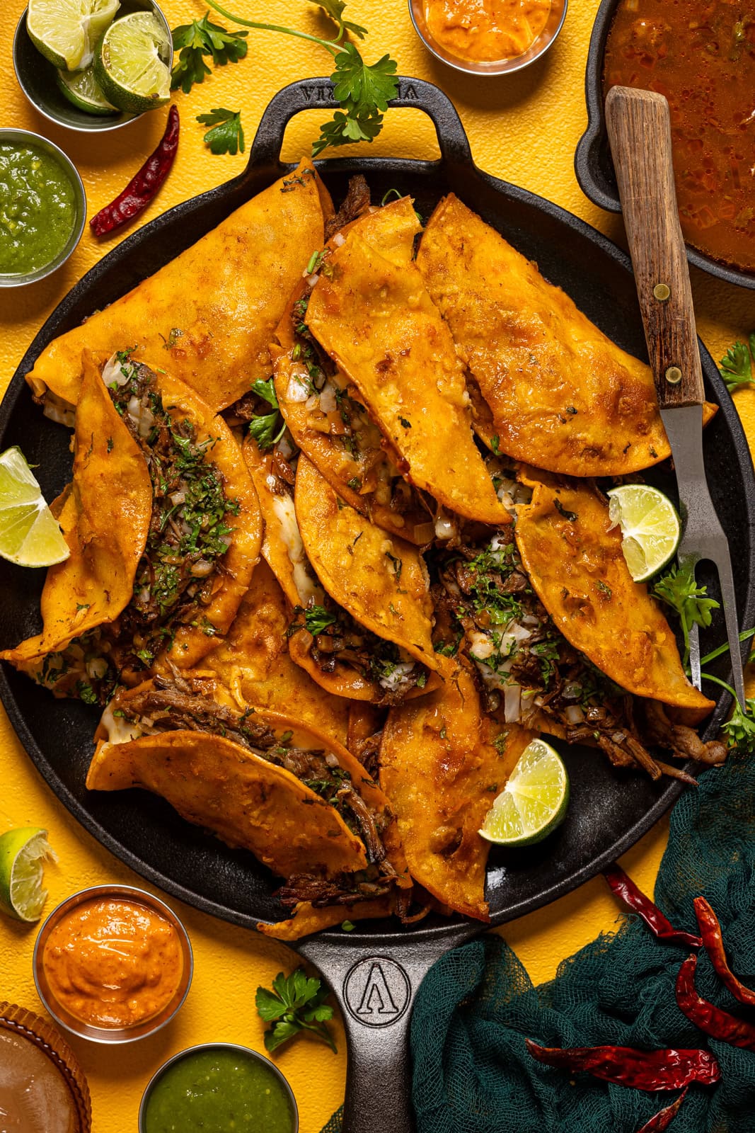 Birria tacos on a platter with a fork, lime, sauces, and peppers on a yellow table.