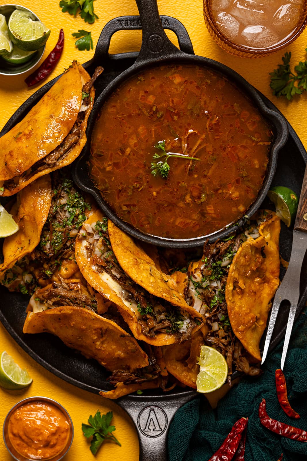 Tacos on a platter with consomme sauce, a drink, dipping sauces, and lime.