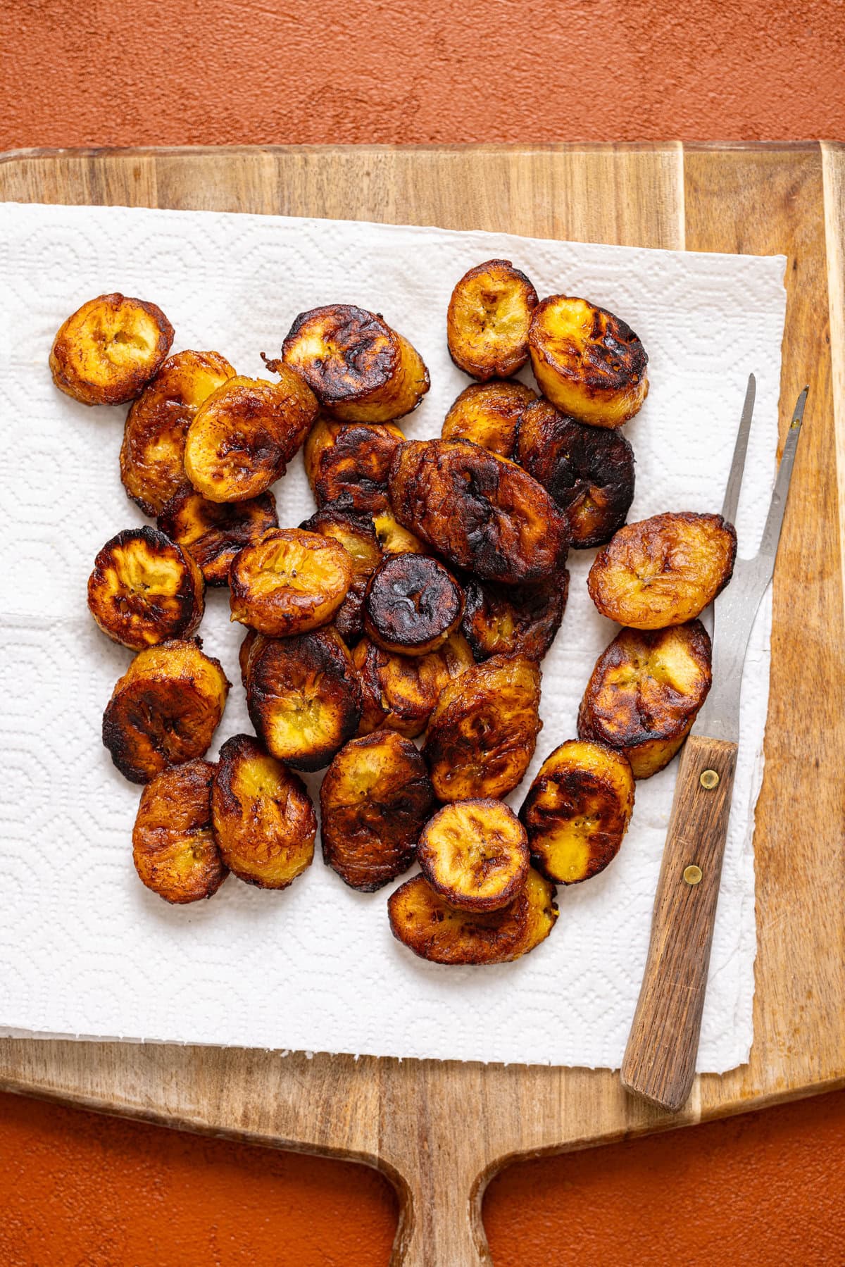 Fried plantain on a paper towel on a cutting board with a fork.