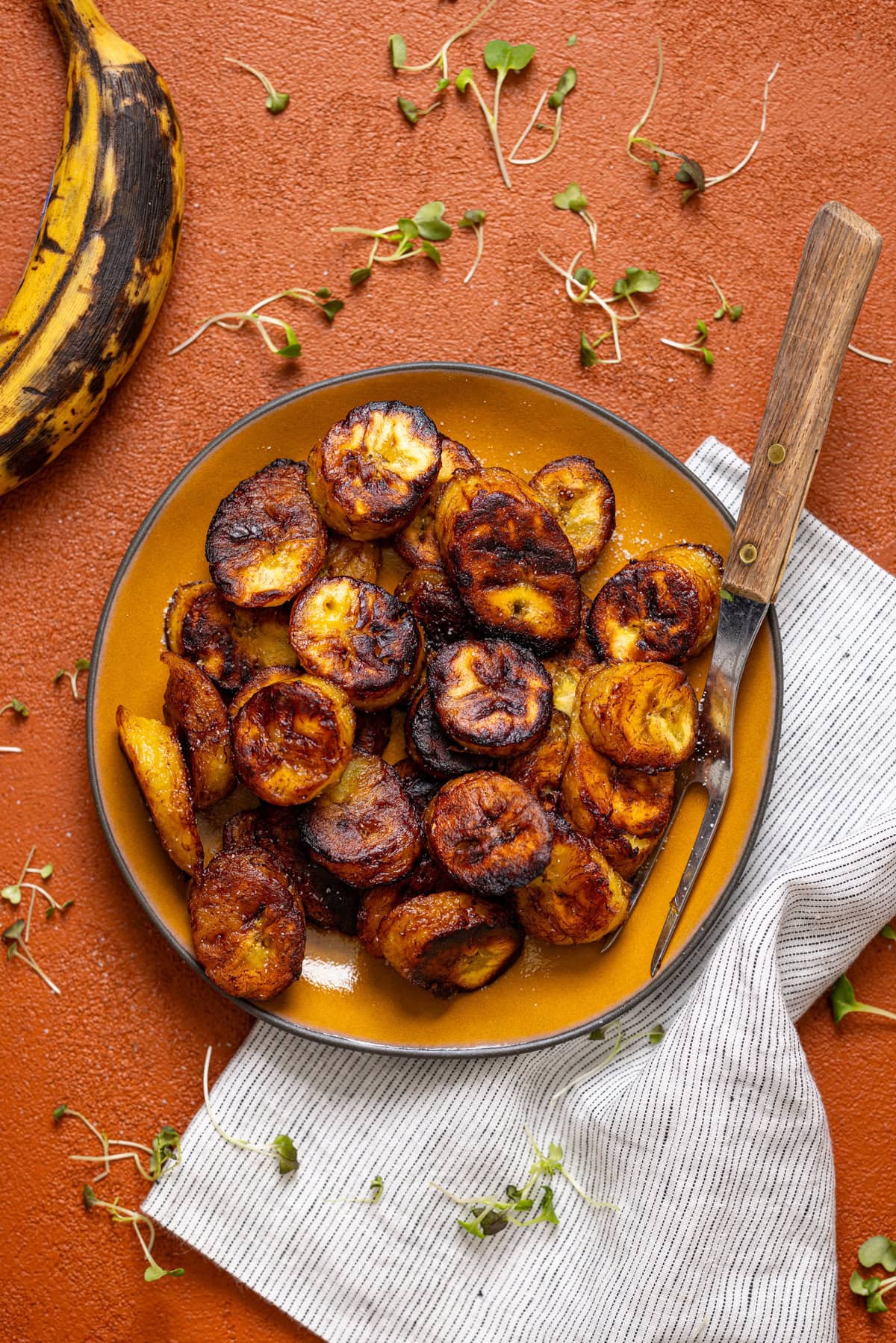 Fried sweet plantain on a yellow plate with a fork, napkin and ripe plantain.