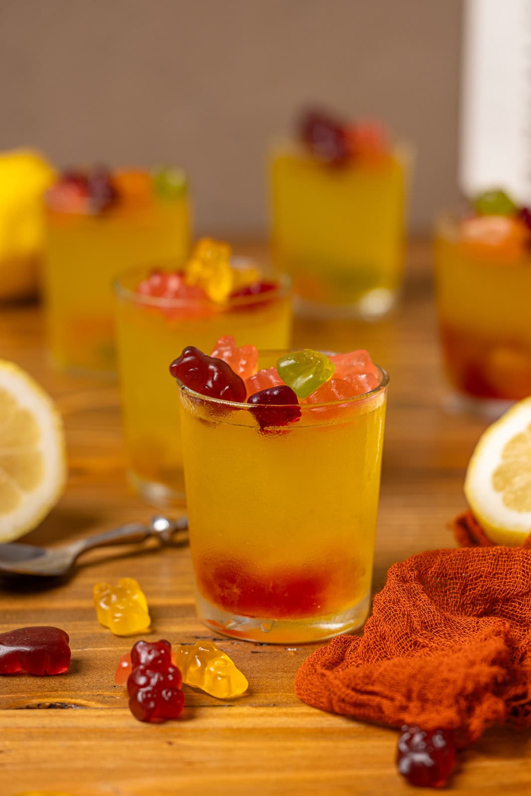 Lemon drop shots lined together on a brown wood table with gummy bears and lemon.