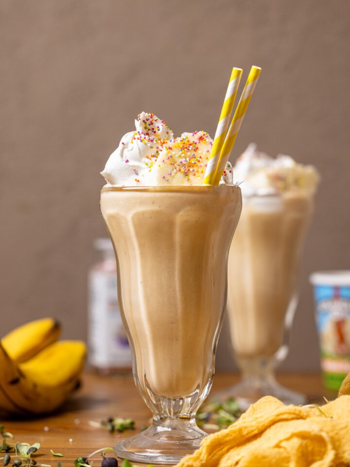 Two glasses filled with milkshake, toppings, bananas, and a yellow napkin.