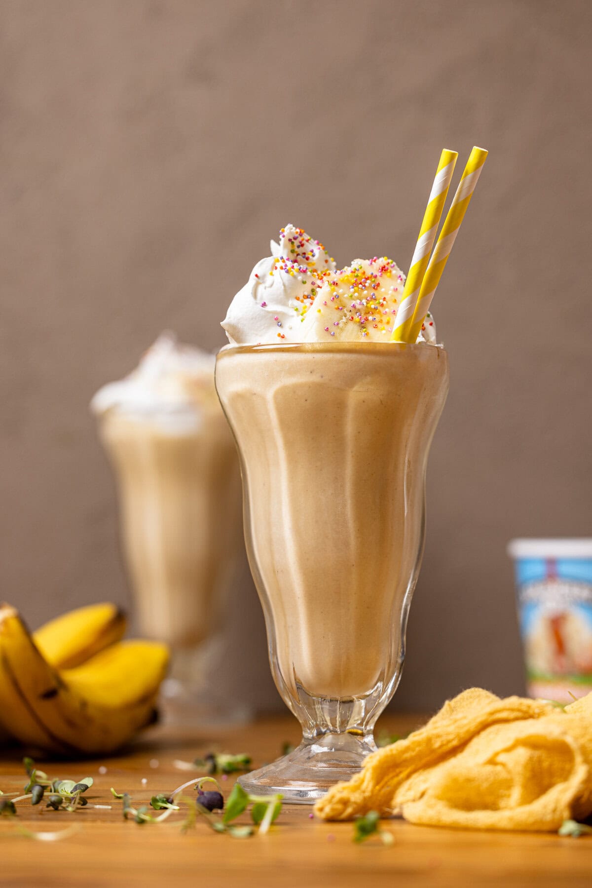 Two glasses of milkshake with yellow strip straws, bananas, and a pint of ice cream in the background. 