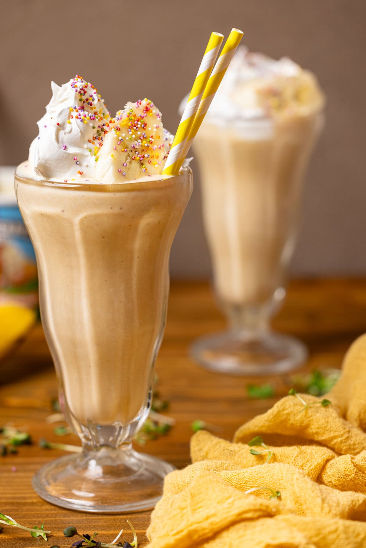 Two glasses of milkshakes with yellow stripe straws, whipped cream, and bananas. 