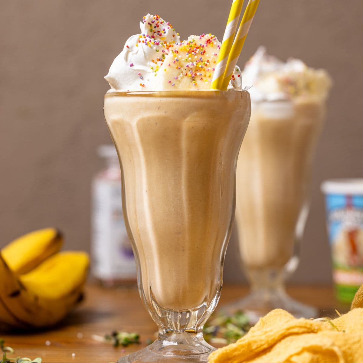 Two glasses filled with milkshake, toppings, bananas, and a yellow napkin.