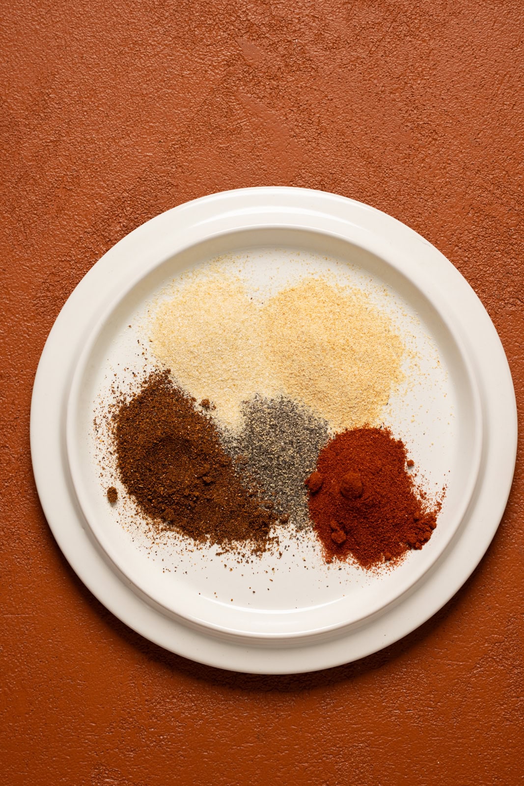 All seasonings grouped together on a white plate on a burnt orange table.