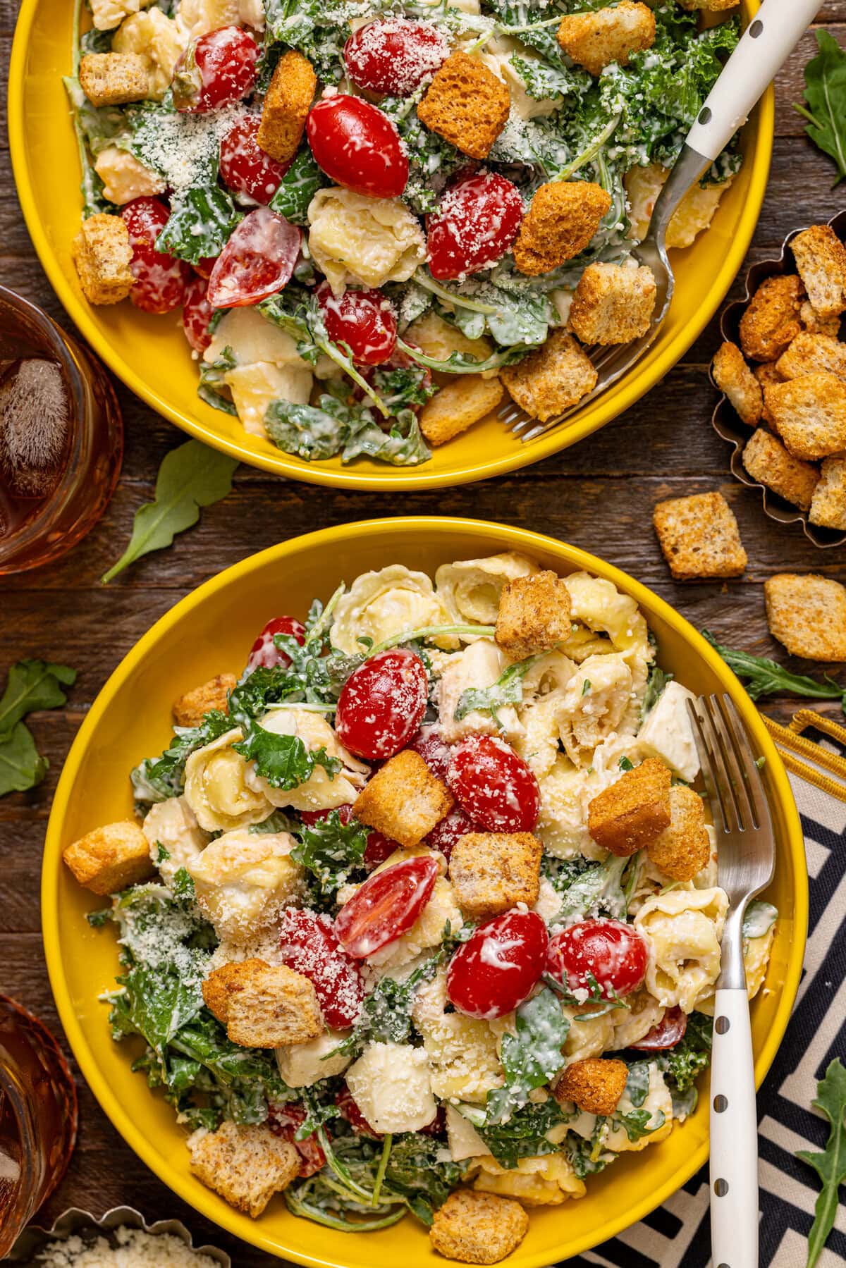 Two bowls of pasta salad with a drink and croutons.