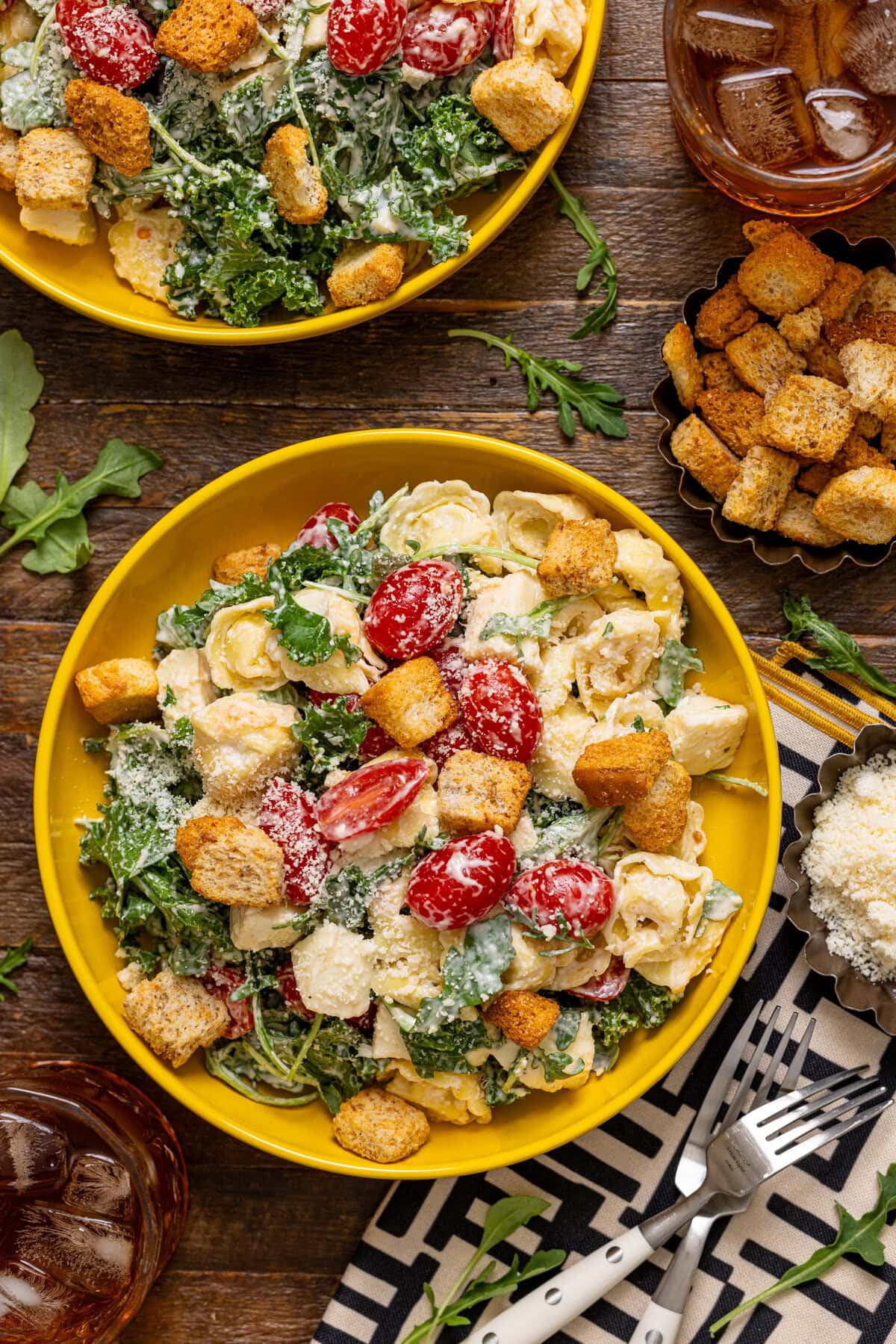 Two bowls of pasta salad on a brown wood table with croutons, a drink, and parmesan.