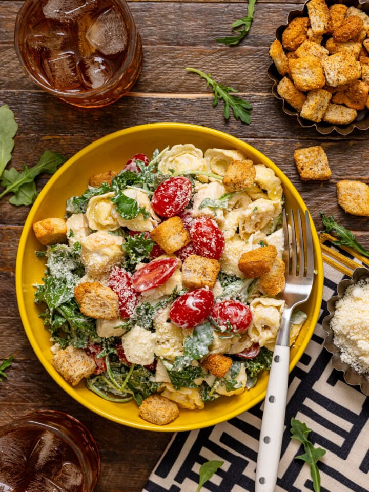 Pasta salad in a yellow bowl on a brown wood table with a fork, drink, and croutons.