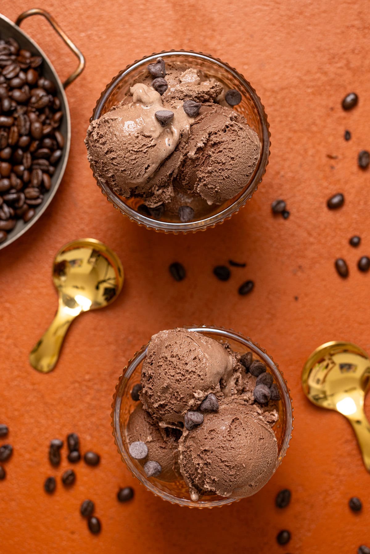 Overview of two bowls of chocolate ice cream on a burnt orange table with two spoons and espresso beans. 