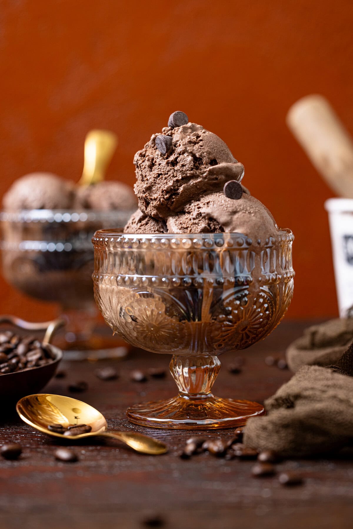Two bowls of chocolate ice cream with a tub of ice cream in the background, spoons, and espresso beans.