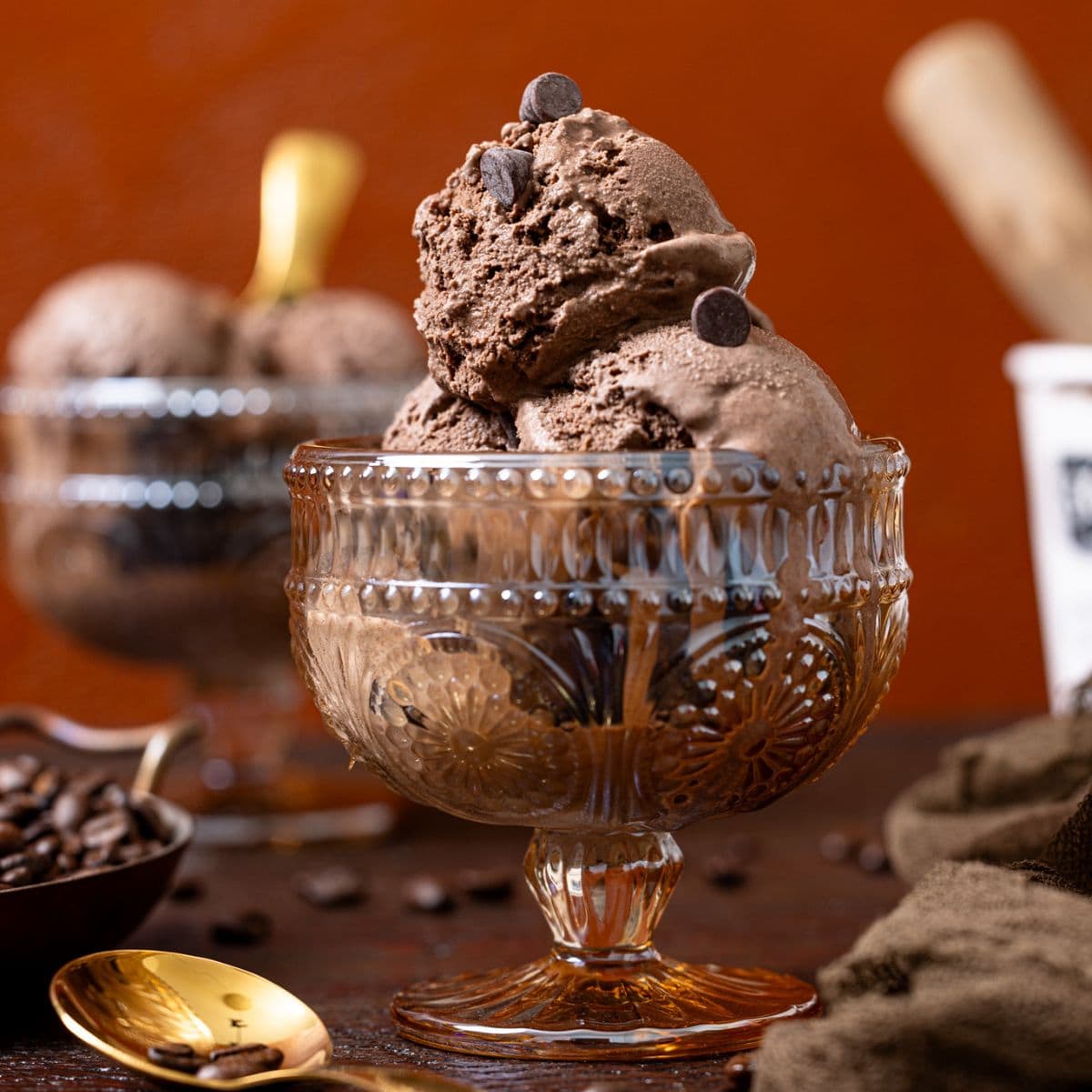 Two bowls of chocolate ice cream with a tub of ice cream in the background, spoons, and espresso beans.