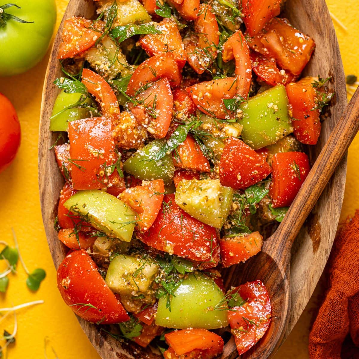 Tomato salad in a wooden serving bowl with a spoon and tomatoes on a yellow table.