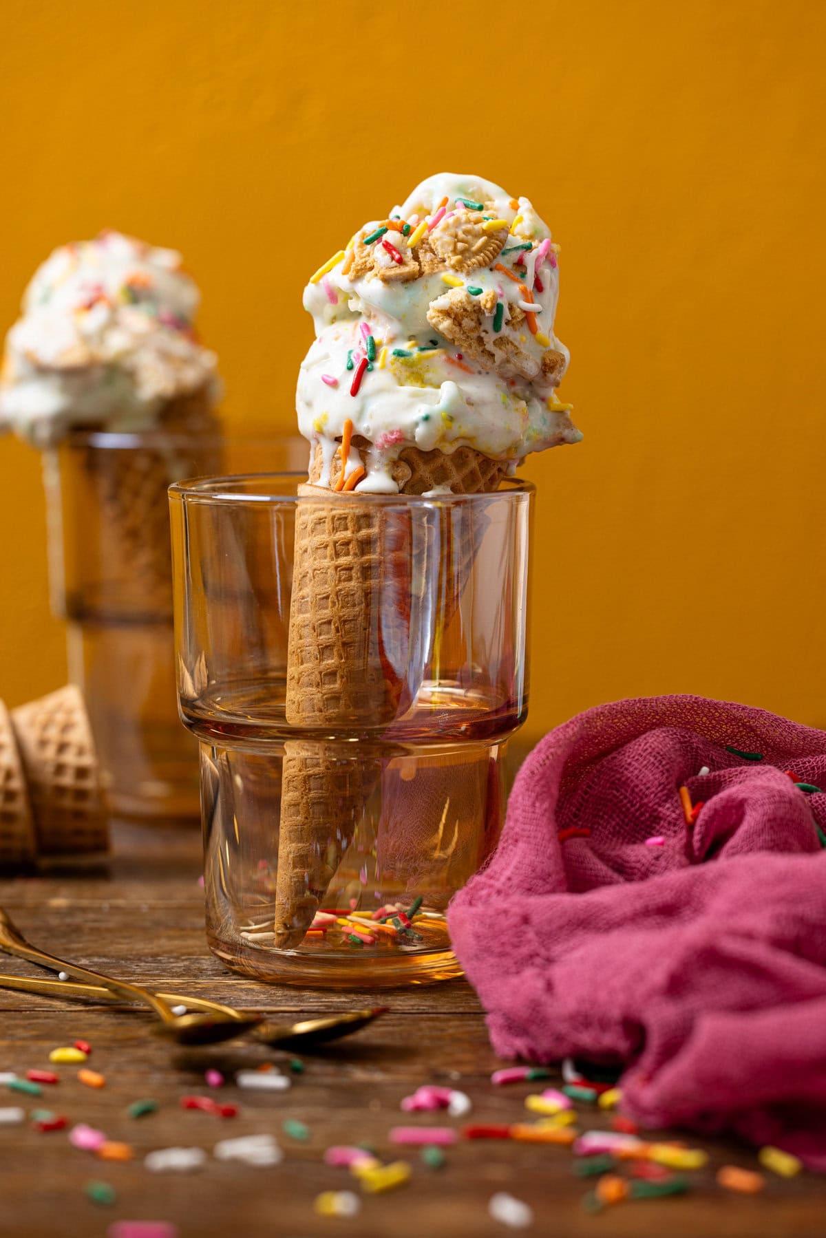 Two ice cream cones in clear glassware with a pink napkin and sprinkles. 