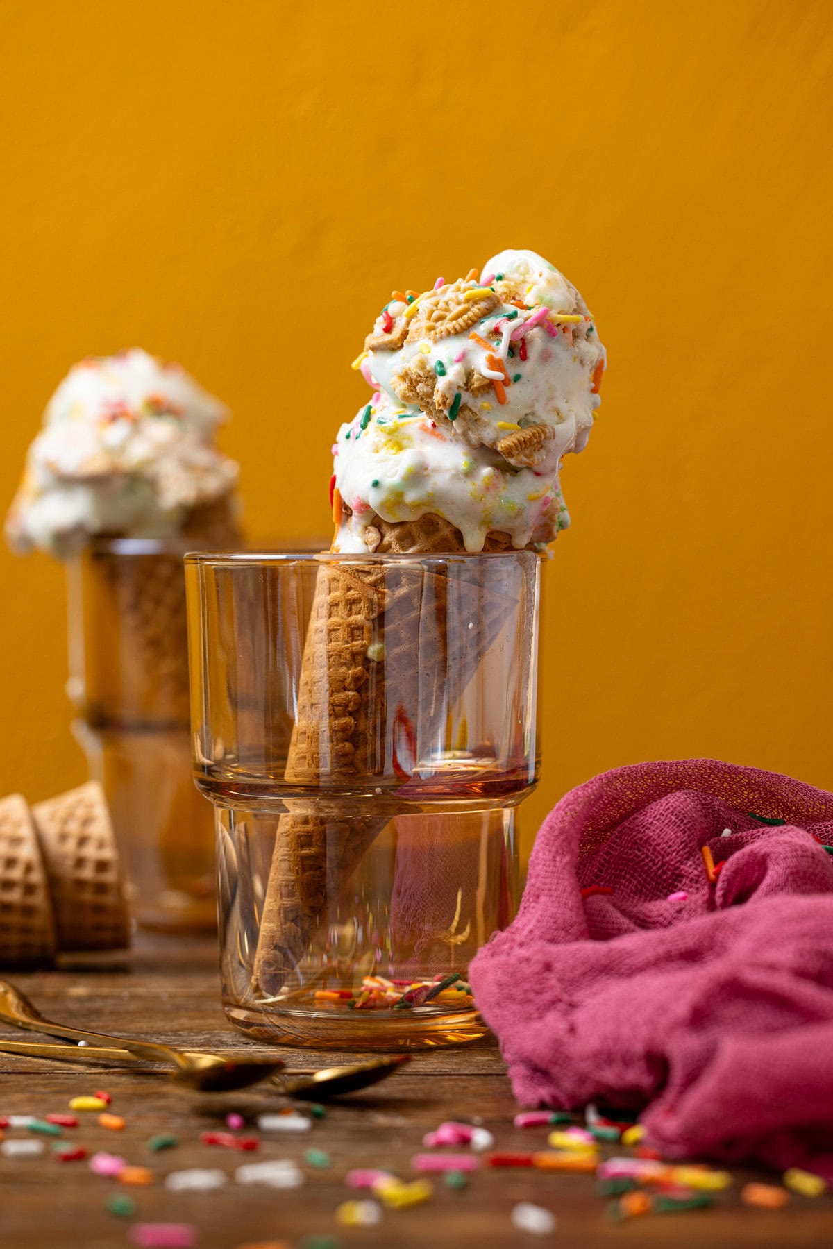 Two ice cream cones in glassware with a pink napkin and cones in the background. 