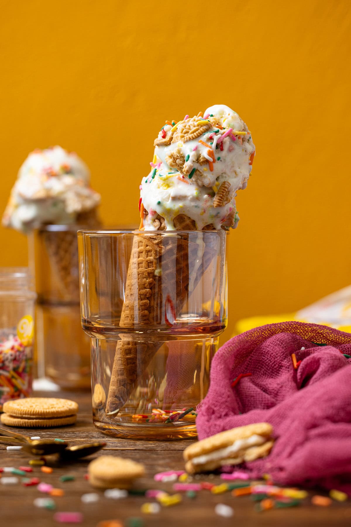 Two ice cream cones in glassware, Oreo cookies, rainbow sprinkles, and pink napkin. 