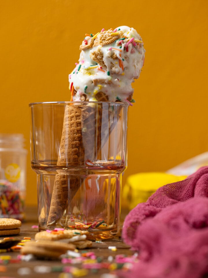 Ice cream cone in glassware in cookies, pink napkin, and sprinkles.