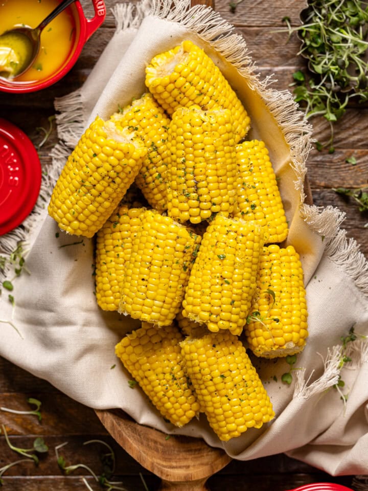 Corn on a wooden platter with melted butter and a spoon and red mini pots.