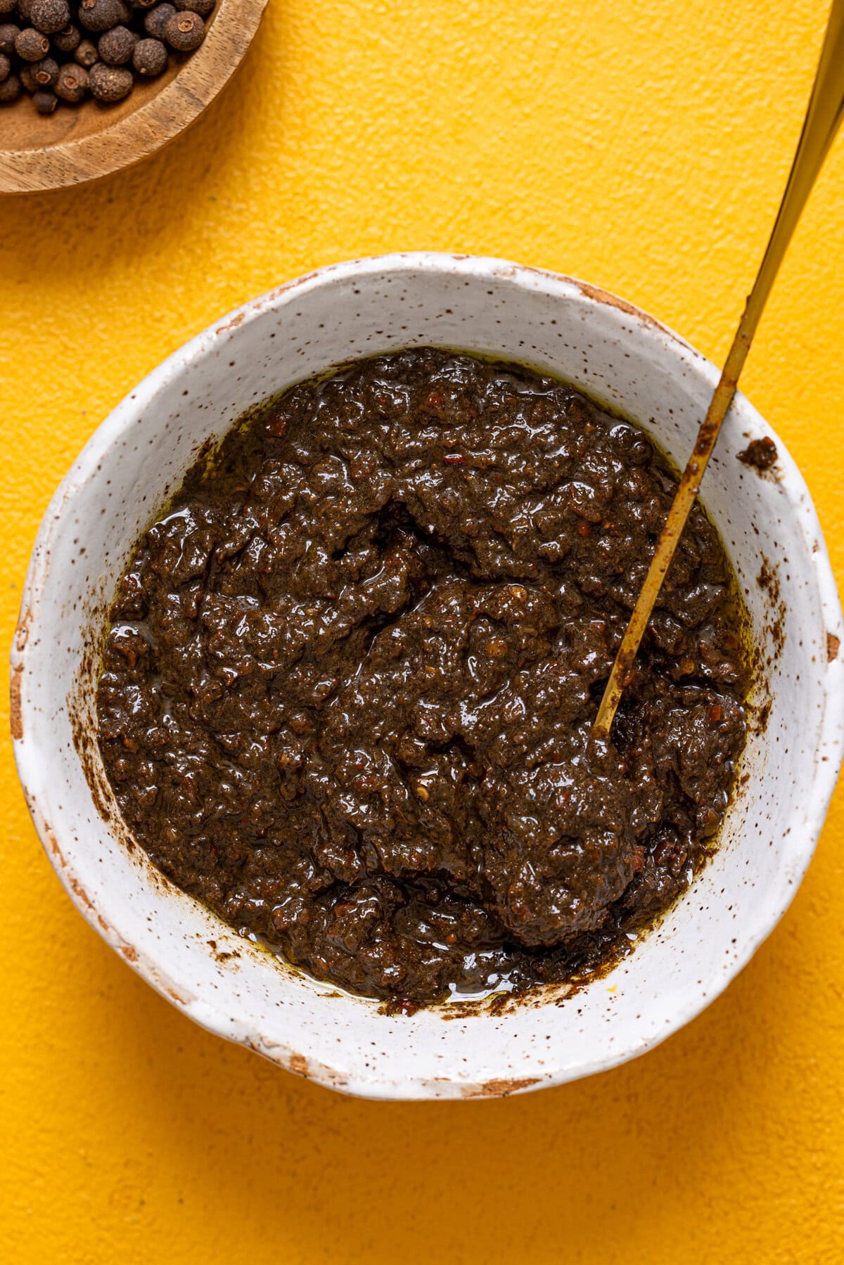 Up close shot of jerk marinade in a white bowl with a spoon on a yellow table.