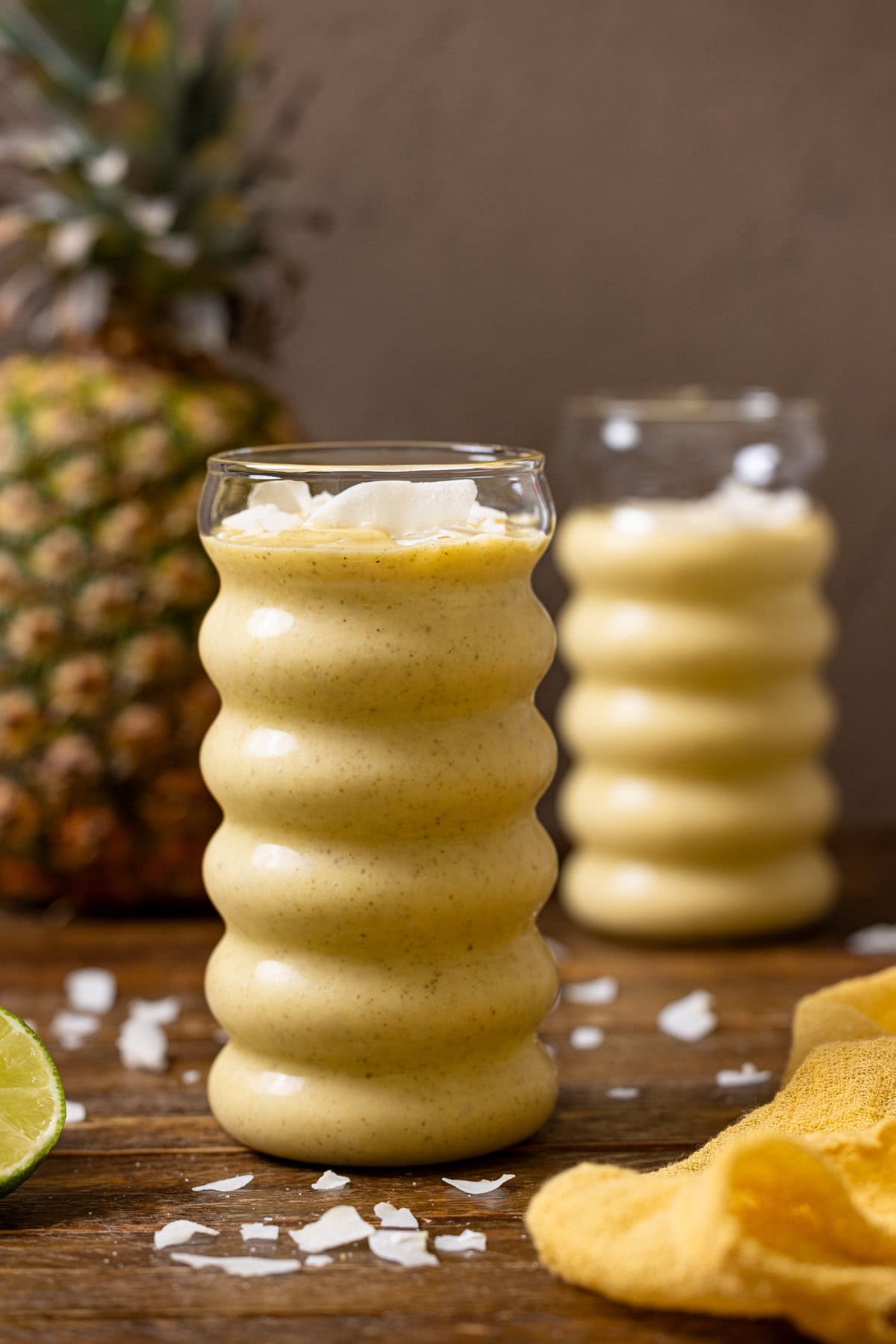 Two glasses of smoothies with a lime, coconut flakes, and pineapple. 