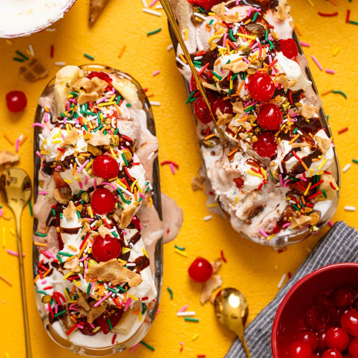 Banana split ice creams on a yellow table with cherries, cones, and more.