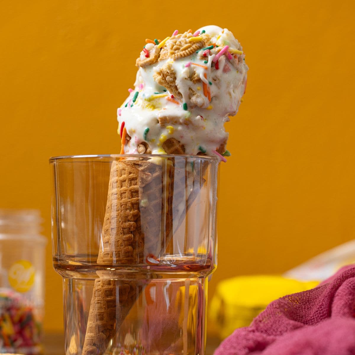 Ice cream cone in glassware in cookies, pink napkin, and sprinkles.